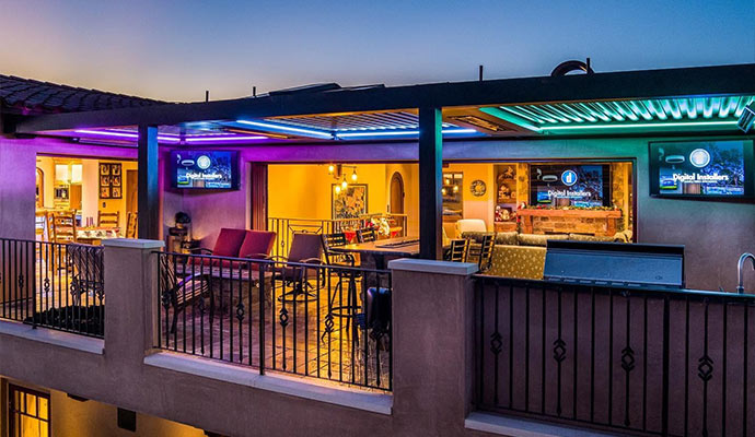 Outdoor rooftop patio showroom with illuminated pergola, bar seating, and outdoor TV screens at night.