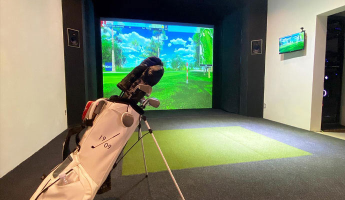 Golf simulator studio with a golf bag and green turf mat in front of a virtual course screen.