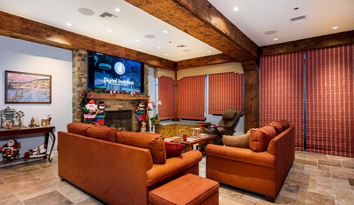 Luxurious rustic living room featuring dark orange velvet sofas, exposed wood ceiling beams, a stone fireplace, and a large central TV display
