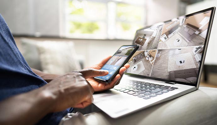 Person using a smartphone to monitor home security cameras on a laptop