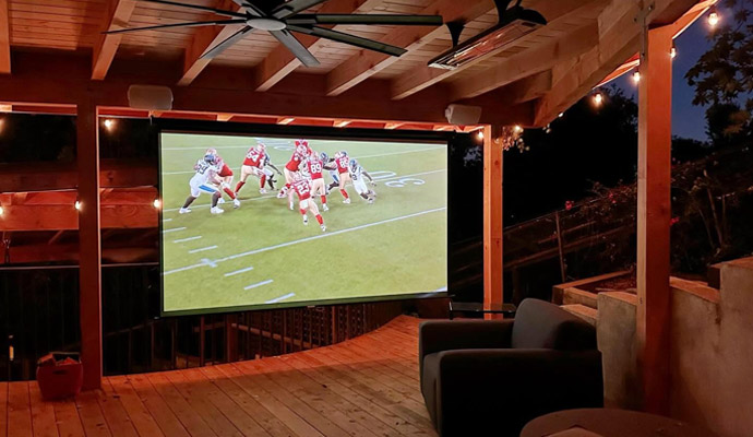 Covered outdoor patio theater with large projection screen showing a football game at dusk.