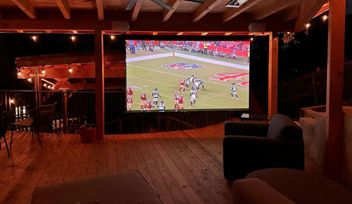 Covered outdoor patio lounge with large projection screen broadcasting a live football game.