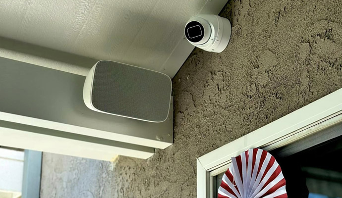 Surface-mounted outdoor audio speaker and security camera installed on an exterior wall.