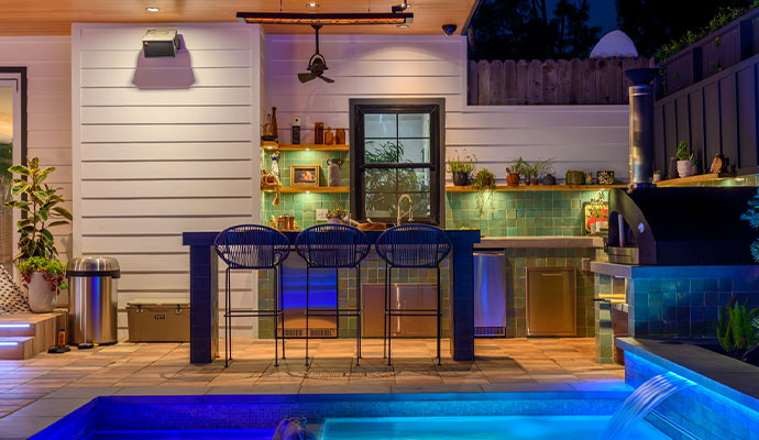 Outdoor kitchen patio by a pool with a wall-mounted speaker.