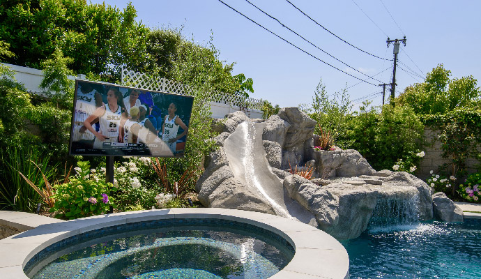 Weatherproof outdoor television screen installed beside a pool rock waterfall and slide.